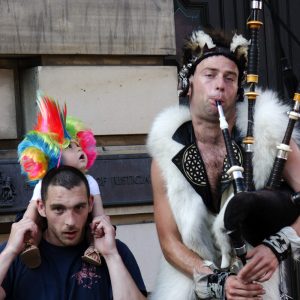 A Edinburgh, un père son enfant sur les épaules et un joueur de cornemuse vêtu d'une peau de bête fixent le photographe. L'enfant une coiffe multicolore sur la tête regarde ébahi la cornemuse, en 2011. Série Regard Léger.